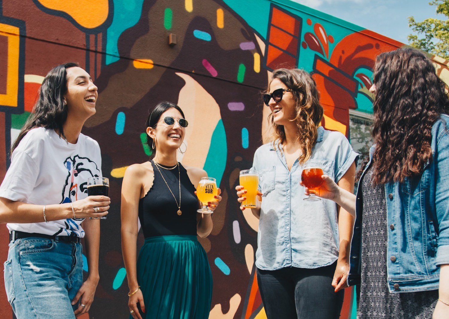 Four friends laughing together outdoors in front of a brightly painted mural