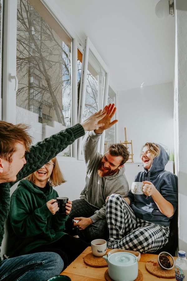 Four friends laughing and high-fiving over mugs of tea in a bright sunlit room